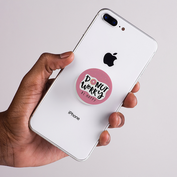 Hand holding a white iPhone with a 'Donut Worry Be Happy' pop socket on a light gray background