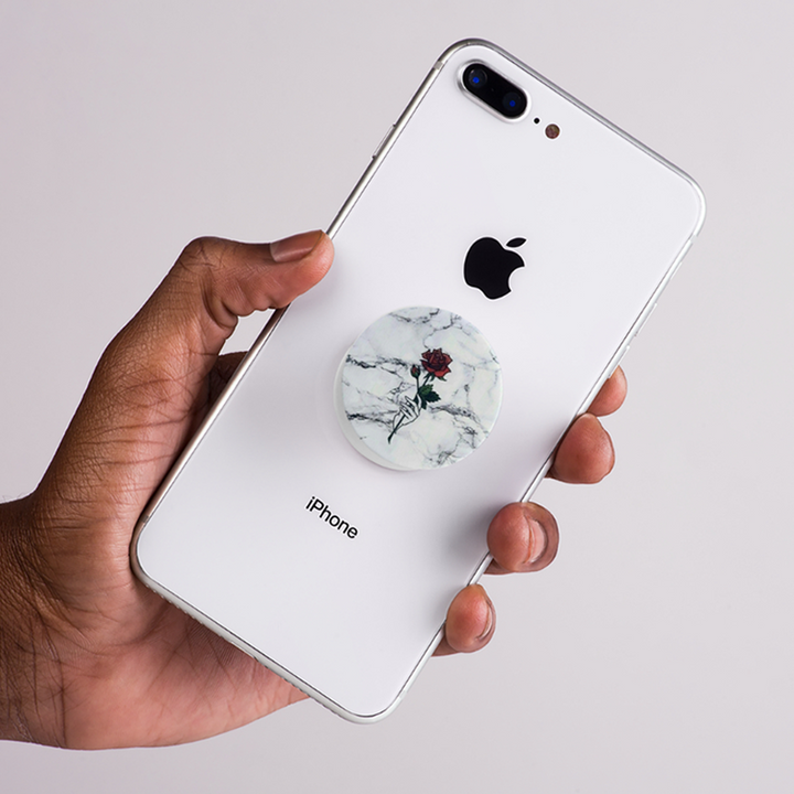 Hand holding an iPhone with a marble-patterned pop socket on a light gray background