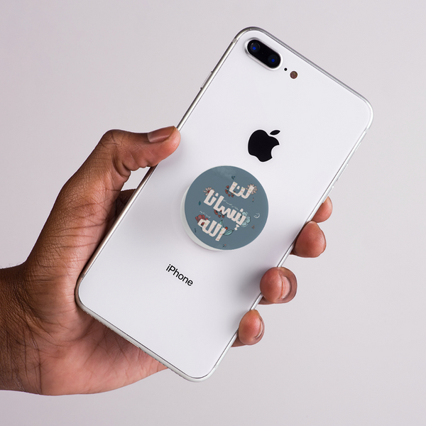Hand holding a white iPhone with a blue popsocket and Arabic text ( lan ynsana allah )on a light gray background